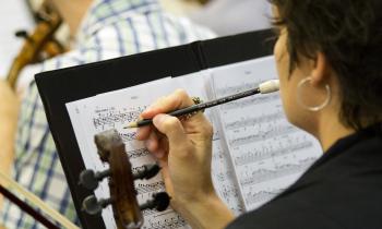 Mulher branca, de cabelos curtos e usando um brinco de argola, segura um lápis enquanto faz anotações em uma partitura. Segura também um violino. Ao fundo, outros músicos. 