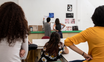 Foto de pessoas que analisam trabalhos artísticos em um ambiente interno. Algumas pessoas estão sentadas ao redor de uma mesa, de costas para a câmera, observando outras duas que estão em pé próximas a uma parede branca. Na parede, estão dispostas diversas obras e trabalhos artísticos. Uma pessoa aponta para um dos trabalhos na parede enquanto a outra observa.