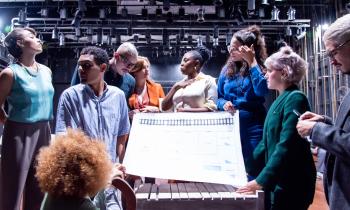 Foto de 9 pessoas, negras e brancas, em um palco de teatro.  Elas estão de pé, em torno de uma grande  folha de papel em que está impressa uma espécie de planta baixa . As pessoas vestem roupas coloridas. Ao fundo, há vários holofotes e uma parede preta.