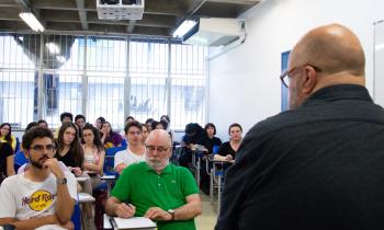 Foto de uma sala de aula. Um homem branco, calvo, usando oculos e blusa preta, está de costas. Um grupo de pessoas, sentadas em carteiras escolares, está a sua frente, e o observa. Ao fundo, janelas e uma parede branca.