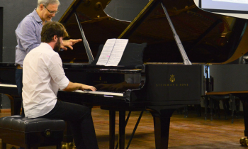Foto de uma aula de piano. Um jovem, de cabelos castanhos e barba, está sentado de costas, com um piano preto a sua frente e tocando o instrumento. Ele veste uma camisa branca, calça preta e tenis. Ao seu lado, está um homem branco, de cabelos curtos e grisalhos, usando óculos e uma camisa azul e branca listrada. Ele observa as suas duas mãos, enquanto conversa com o jovem. O chão é de madeira e ao fundo uma parede preta e a vista parcial de um projetor