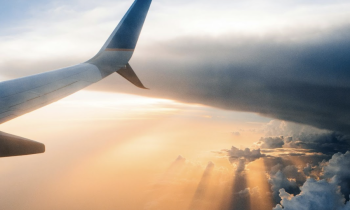 Foto da asa direita de um avião. A asa está posicionada no canto esquerdo da foto. No horizonte há nuvens contornadas por diferentes tons de laranja e sombreados de um sol poente.
