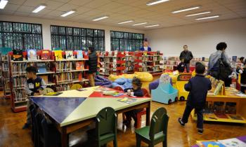 Foto de sala de grupo de crianças e adultos espalhados em uma sala de biblioteca. Em primeiro plano, crianças brincam sentadas em uma mesa próxima, no fundo, há várias estantes de livros. 