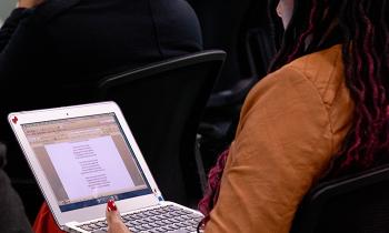 Foto de uma mulher negra lendo um texto em um laptop em seu colo. Ela tem dreads vermelhos e compridos, usa blazer caramelo e unhas vermelhas e está sentada de costas para a câmera, em um auditório. À frente dela, parte de uma cadeira e de uma pessoa de costas.