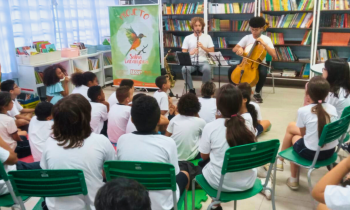 Foto de uma sala de aula, em que um grupo de cerca de 20 crianças - sentadas em cadeiras verdes - assistem a apresentação musical. As crianças estão de costas e vestem camisetas brancas e bermudas azuis-marinho. À frente delas, estão dois jovens, ambos sentados, um deles tocando clarinete e o outro, violoncelo. Ao fundo, uma estante cheia de livros.