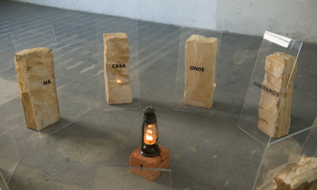 Foto de lampião aceso sobre um pedestal com diversas pedras em semicírculo ao seu redor. As pedras são claras e na frente de cada uma há uma placa transparente com os dizeres, da esquerda para a direita: “na”, “casa”, “onde”, “moramos”. O fundo é cinza claro. 