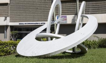 Foto de uma escultura branca, formada por arcos circulares. Ela está localizada em um gramado verde. Ao fundo, um edifício na cor bege