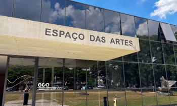 Foto de um edifício com janelas de vidro escuro espelhado e um letreiro escrito Espaço das Artes em uma estrutura de concreto que serve de cobertura sobre a entrada. Acima do edifício, o céu está azul com algumas nuvens brancas. O reflexo dos vidros mostra grama e árvores verdes, carros, pessoas e outros prédios. 