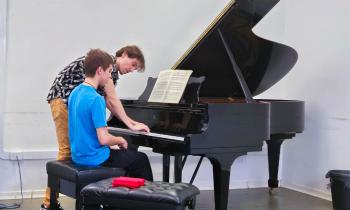 Foto de dois homens brancos jovens em frente a um piano de cauda preto. Ambos têm cabelos curtos, lisos e castanhos claros. Um deles, de camiseta azul e calça preta, está sentado em um banco preto em frente ao piano. O outro, de camiseta preta e branca e calça bege, está em pé tocando o piano. Sobre o piano, uma partitura. As paredes são brancas e, do lado deles, outro banco preto sobre o qual há um tecido vermelho.  