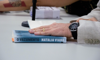 Foto de uma mão de pessoa branca repousando sobre um livro azul que está sobre uma mesa clara. O livro está com um marcador laranja e tem escrito na sua lombada “O Vazamento” e “Natalia Viana”. A mão tem uma aliança dourada no dedo anelar, um relógio preto no pulso e uma manga cinza de paletó com botões. Ao fundo e desfocada, a base de um microfone, uma caixa branca e o braço de outra pessoa branca. 