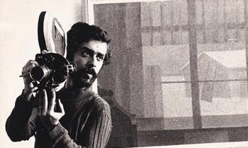 Foto em preto e branco de homem branco de cabelos e barba escuros segurando uma câmera de cinema apoiada sobre seu ombro. Ele usa camisa de manga comprida e escura. Ao fundo, um quadro abstrato.