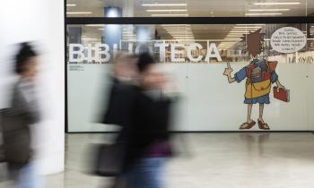 Foto da entrada da biblioteca da ECA. Sobre uma divisória de vidro, destaca-se em letras grandes e brancas a palavra ‘biblioteca’ e uma ilustração do cartunista Dorinho de uma pessoa de costas com uma mochila repleta de livros. Em primeiro plano e em desfoque, três pessoas caminham pelo saguão. 