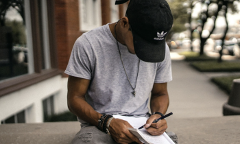 Foto de um jovem sentado em uma mureta de concreto escrevendo em um caderno apoiado no colo. Ele é negro, usa camisita cinza, calça jeans cinza, colar, pulseiras, relógio e um boné preto com logo da adidas. Ele está olhando para baixo, com a aba do boné cobrindo o rosto. O fundo da imagem está desfocado, com um prédio de tijolos vermelhos, janelas brancas e árvores na calçada.
