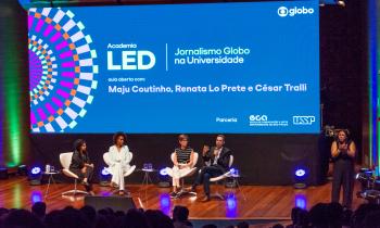 Foto de cinco pessoas em um palco diante de uma plateia. Quatro delas, duas mulheres negras, uma mulher branca e um homem, estão sentadas em poltronas brancas. O homem fala ao microfone e gesticula olhando para a plateia, enquanto as mulheres o olham. No canto direito do palco, uma mulher negra está de pé fazendo linguagem de sinais. No fundo do palco, um grande telão exibe os dizeres ‘Academia LED. Jornalismo Globo na Universidade. Aula aberta com Maju Coutinho, Renata Lo Prete e César Tralli. 