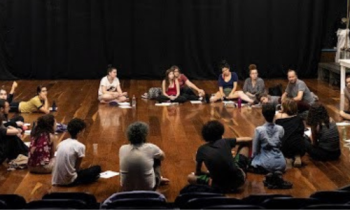 Foto de 23 pessoas sentadas em roda em um palco de teatro com piso de madeira. Elas vestem roupas coloridas. Ao fundo uma cortina preta e do lado direito da imagem há uma mesa, um manequim e cordas. No primeiro plano da imagem, as cadeiras pretas da plateia. 
