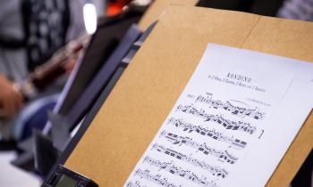 Foto de uma partitura impressa em uma folha de papel apoiada sobre um suporte de madeira. Ao lado dela, há um metrônomo digital e ao fundo há a silhueta de uma pessoa tocando um fagote.