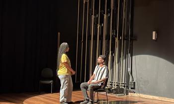 Foto de duas pessoas em um palco interagindo. Uma mulher negra com tranças longas e pretas, camiseta amarela e calça clara e tênis preto, está em pé, de frente para um homem sentado em uma cadeira. Ele é negro, tem cabelos curtos pretos, camiseta de listras verticais pretas e brancas, de calça cinza e tênis pretos. Há foco de luz sobre os dois, o chão é de madeira e a parede ao fundo é preta com cordas.