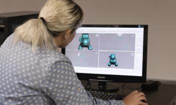 Foto de pessoa branca com cabelos loiros presos em um rabo de cavalo e vestindo uma camisa azul-clara de bolinhas brancas operando um computador com um mouse. Ela está sentada, de costas para a câmera, usando um software de modelagem 3D, que exibe um modelo tridimensional de um personagem ou objeto com aparência de boneco no monitor. 