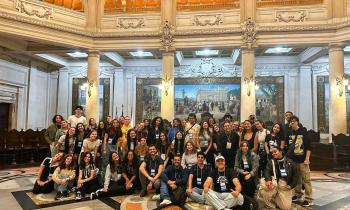 Foto de um grupo de cerca de 50 pessoas, majoritariamente mulheres jovens e brancas, todos com crachás pendurados no pescoço, dentro de um edifício antigo. O piso é feito de desenhos geométricos em mármore cinza, preto e laranja. Atrás das pessoas, colunas com luminárias e uma pintura grande pendurada na parede. O teto também é construído de abóbodas e desenhos esculpidos. 