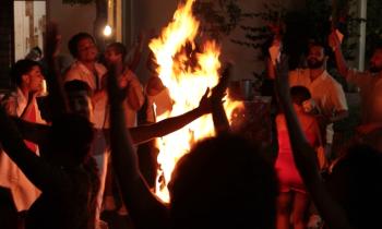 Foto de grupo de pessoas em um círculo em volta de uma fogueira. A silhueta das pessoas em primeiro plano é escura, enquanto as mais distantes, iluminadas pelo fogo têm tons avermelhados. O fundo é escuro.