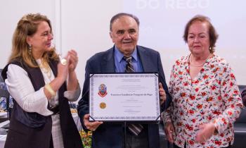 Foto de três pessoas em pé, lado a lado. No centro, um homem branco, idoso, vestindo terno escuro, camisa azul e gravata listrada, segurando e exibindo um certificado. À esquerda, uma mulher branca de cabelos loiros e longos, vestida com uma roupa preta e branca, está batendo palmas. À direita, há outra mulher branca, idosa, de cabelos castanhos curtos, vestindo uma blusa florida. Ao fundo, tela de projeção exibindo texto em azul e branco.