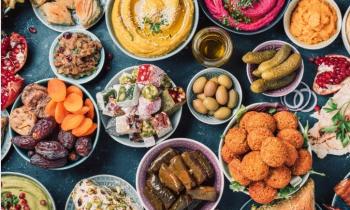 Foto a partir de cima de uma superfície azul sobre a qual estão cerca de 20 recipientes com diferentes alimentos muito coloridos: frutas secas, pastas, bolinhos fritos, pepinos, romãs, pães, entre outros. 