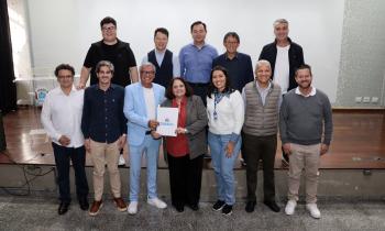 Foto de um grupo de 12 pessoas, de pé e posicionadas em duas fileiras. Sete pessoas à frente e cinco pessoas atrás, em cima de um palco de madeira, em sua maioria homens brancos. Ao centro, um homem negro, de cabelos brancos e curtos vestindo um terno azul claro e uma mulher parda, de cabelos de tamanho médio, vestindo um terno cinza e uma camisa vermelha, seguram e exibem uma pasta branca com a palavra “Suzano” impressa em azul. Ao fundo, uma tela branca e o piso de madeira.