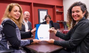 Foto de duas mulheres - ambas brancas, uma de cabelos longos e loiros e outra de cabelos castanhos cacheados - sorrindo enquanto posam segurando um documento com capa azul e branca, com os logotipos "eca" e "USP" em azul. Atrás delas, um homem branco careca e com barba e uma mulher amarela de cabelos castanhos e curtos também sorriem. Todos estão sentados em torno de uma mesa retangular de madeira. Ao fundo, uma parede vermelha com prateleiras embutidas e um quadro.