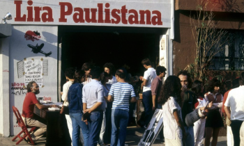 Foto da fachada do espaço cultural Lira Paulistana com uma multidão reunida na calçada. Pessoas conversam e folheiam livros, enquanto um homem sentado a uma mesa vermelha autografa obras. O letreiro “Lira Paulistana” está em destaque acima da entrada.