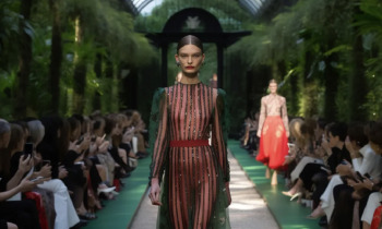 Mulher branca caminha por uma passarela cercada por vegetação verde em um desfile de moda. Ela veste um vestido longo verde e transparente com listras verticais em vermelho e preto, com detalhes brilhantes. Ela tem cabelos castanhos lisos que estão presos. Usa botas, cinto e batom vermelhos. Várias pessoas estão sentadas em ambos os lados da passarela, tirando fotos com celulares. No fundo desfocado, outra mulher branca se aproxima usando uma saia vermelha. 