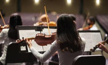 Foto de  músicos em uma apresentação ou ensaio orquestral, vistos por trás. O foco está em uma violinista de cabelos escuros, que segura seu violino e arco enquanto lê uma partitura apoiada em um suporte à sua frente. Ela veste uma blusa clara e está sentada em uma cadeira preta. Ao redor dela, outros músicos também tocam instrumentos de corda, como violinos e violas. Ao fundo, uma plateia desfocada, com assentos vazios.