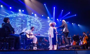 Foto de um palco durante um concerto. No centro, uma mulher negra, de cabelos crespos e loiros, está em pé diante de um microfone, usando um vestido escuro e um casaco branco e longo. Ao lado dela, um maestro de cabelos brancos usando um terno gesticula com uma das mãos erguidas, de frente para a orquestra. Atrás da cantora, um músico de roupas claras toca um violão sentado. À esquerda e à direita, membros da orquestra estão sentados com violinos, violoncelos e outros instrumentos.