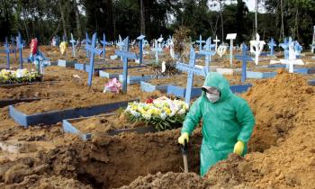 Foto de um cemitério com diversos túmulos demarcados com estruturas de madeira simples com cruzes azuis e brancas sobre a terra. Em alguns deles há flores. Em primeiro plano, há uma pessoa em pé dentro de uma cova segurando uma pá apoiada ao chão. Ela veste uma capa de plástico verde com capuz, boné, máscara e luvas de borracha amarelas. Ao fundo, árvores.