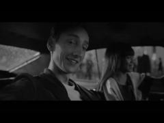 imagem em preto e branco de campanha publicitária em que aparecem um jovem branco, que sorri e olha para a camera, e ao fundo uma mulher, levemente desfocada, que também sorri enquanto olha a frente. eles estão dentro de um carro.