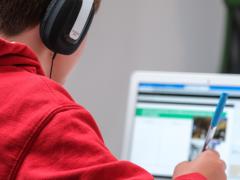 Foto de um garoto escrevendo, enquanto usa um laptop e fones de ouvido. O menino, que é branco e possui cabelo escuro, usa uma blusa de moletom vermelha e fones de ouvido pretos e cinzas. Ele segura com a mão direita uma caneta transparente de tampa azul. Ao fundo, há um laptop branco com a tela desfocada. Em último plano, está uma parede branca.