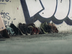 Foto de diversos pares de sapatos gastos encostados em uma parede. A parede apresenta alguns traços de grafite. A foto foi tirada na altura do chão.