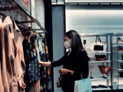 Foto de uma mulher escolhendo roupas em uma loja. A mulher é branca, possui cabelos lisos e escuros e está usando máscara. A roupa dela é preta e ela está com um relógio no braço esquerdo, no qual pendura uma sacola de compras. Essa mulher está escolhendo roupas entre várias opções penduradas em uma arara. Ao fundo, pode-se ver prateleiras com bolsas e sapatos.