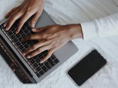 Foto vista por cima de um laptop, um celular, e mãos de uma pessoa. Tudo está sobre um lençol branco amassado. As mãos, que são de uma pessoa que está usando uma blusa branca até os pulsos, estão sobre as teclas do laptop. Ao lado do laptop encontra-se o celular.