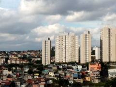 Foto de um bairro da região leste de Osasco. É possível observar uma comunidade composta por casas de baixo padrão cercada por prédios residenciais de classe média. O céu, ao fundo, tem muitas nuvens.