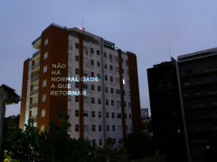foto de um fim de tarde nublado, de um prédio branco e marrom. Nele, está projetada em luz a frase não há normalidade a que retornar.