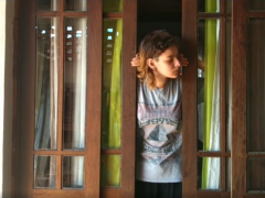 mulher jovem, branca, de cabelos lisos, castanhos e na altura do ombro, abra com as duas mãos uma porta de madeira e vidro. Ela veste uma camiseta cinza, estampada e olha para o lado direito.