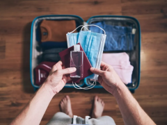 ao centro da imagem, duas mãos seguram duas máscaras, um frasco de alcool em gel e um passaporte. Ao fundo, uma mala aberta com roupas dobradas