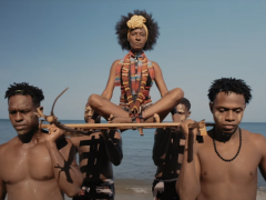 Foto mostra uma mulher jovem e negra de cabelos escuros e crespos em um corte redondo, como uma coroa. Ela usa um adorno de tecido no cabelo, lembrando uma tiara com flor, e roupas com grafismos geométricos em tons de azul, vermelho e amarelo. Ela está sentada em posição de lótus sobre uma espécie de maca, que é carregada nos ombros por quatro homens negros e jovens. Ao fundo, o mar e o céu, como se as pessoas estivessem saindo da água.