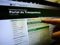 Foto da tela de um computador. Na tela, o site do Portal da Transparência do governo federal. O site possui colunas em cores verde claro e escuro. No canto direito da imagem aparece uma mão com o dedo indicador apontando para a tela. 