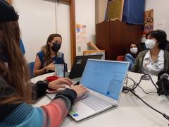 Foto mostra estudantes mulheres sentadas em volta de uma mesa encabeçada por Lilian Viana, uma mulher branca de cabelos castanhos com pontas claras na altura do peito. Ela tem um computador à sua frente e gesticula enquanto fala. Ao seu lado, em primeiro plano, uma estudante digita em um computador. Todas usam máscaras. No fundo, há alguns cartazes e materiais de papelaria.