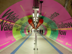 Foto tirada na ponta de um corredor no metrô de São Paulo . As paredes estão totalmente cobertas por cartazes publicitários chamativos (predominantemente em cor-de-rosa e em dois tons de verde) com títulos de músicas famosas escritas em preto. O corredor é bem iluminado. Nele, algumas barras de segurança separam as pessoas que andam nos dois sentidos.