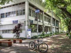 Foto do prédio do CTR. Em primeiro plano, há uma placa azul onde está escrito ECA em branco. No poste que sustenta a placa está amarrada uma bicicleta. Na parte esquerda existem três bancos laranja escuro, sem encosto. Mais ao fundo localiza-se o prédio, que foi fotografado de lado, de modo que é possível ver os dois lados dele. Na frente do prédio há placas em azul identificando o nome e a sigla do departamento. Ao seu lado encontra-se um caminho, ladeado de grama, arbustos e árvores. 