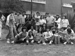 Foto em preto e branco mostra diversos jovens, homens e mulheres, no gramado em frente à fachada do prédio principal da ECA. Alguns estão sentados, enquanto outros estão atrás, de pé. A maioria sorri posando para a câmera e abraçando os colegas, enquanto outros estão distraídos no momento da foto. Alguns ainda fazem brincadeiras com os colegas. 
