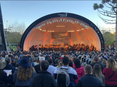 Foto de uma platéia assistindo a apresentação de uma orquestra sinfônica em um palco ao ar livre. Todos os assentos parecem estar ocupados, de modo que algumas pessoas ficam de pé no fundo. À frente do público está um palco com cobertura em meia-lua, no qual se posiciona a orquestra. A iluminação fica entre o laranja e o amarelo. No lado esquerdo do palco, encontra-se uma araucária. No direito, uma estrutura preta que sustenta o sistema de som. 
