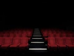 Foto de sala de cinema vazia. Ao centro, há um corredor de escadas. Várias fileiras de poltronas vermelhas podem ser vistas, todas elas vazias. O fundo da imagem é escuro, uma sombra.
